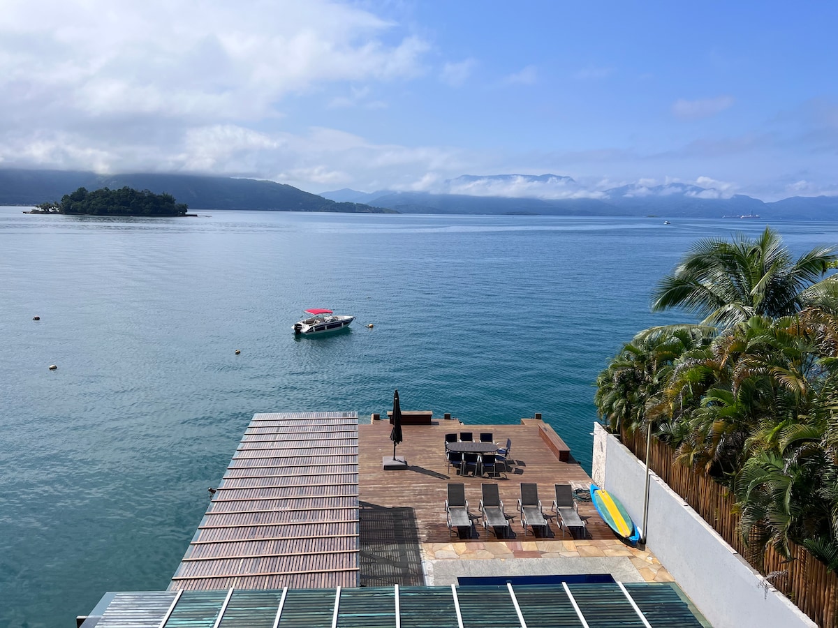 A serene waterfront view is presented, featuring a spacious deck with outdoor seating and lounge chairs. Gentle waves lap against the shore, and a small boat is anchored nearby. Lush palm trees frame the area, enhancing the tranquil coastal atmosphere.