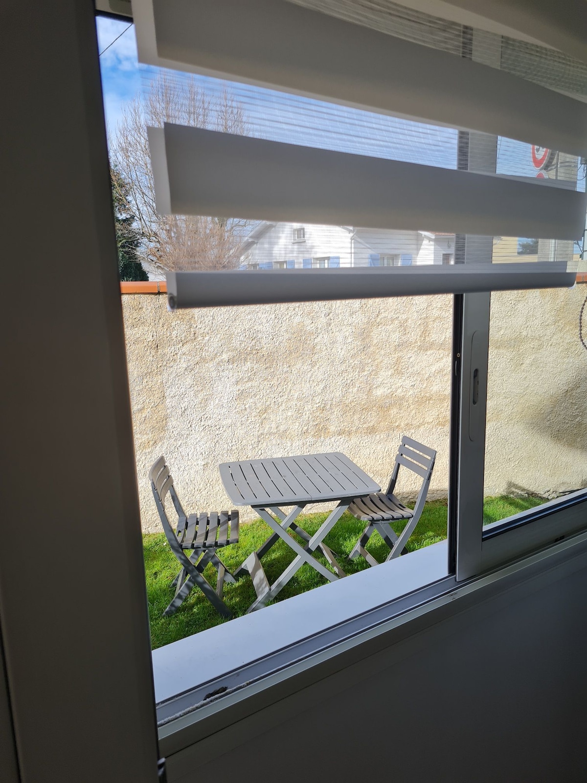 A view from the window reveals a small outdoor terrace featuring a wooden table and two folding chairs positioned on a patch of green grass. A low wall encloses the space, providing a sense of privacy.