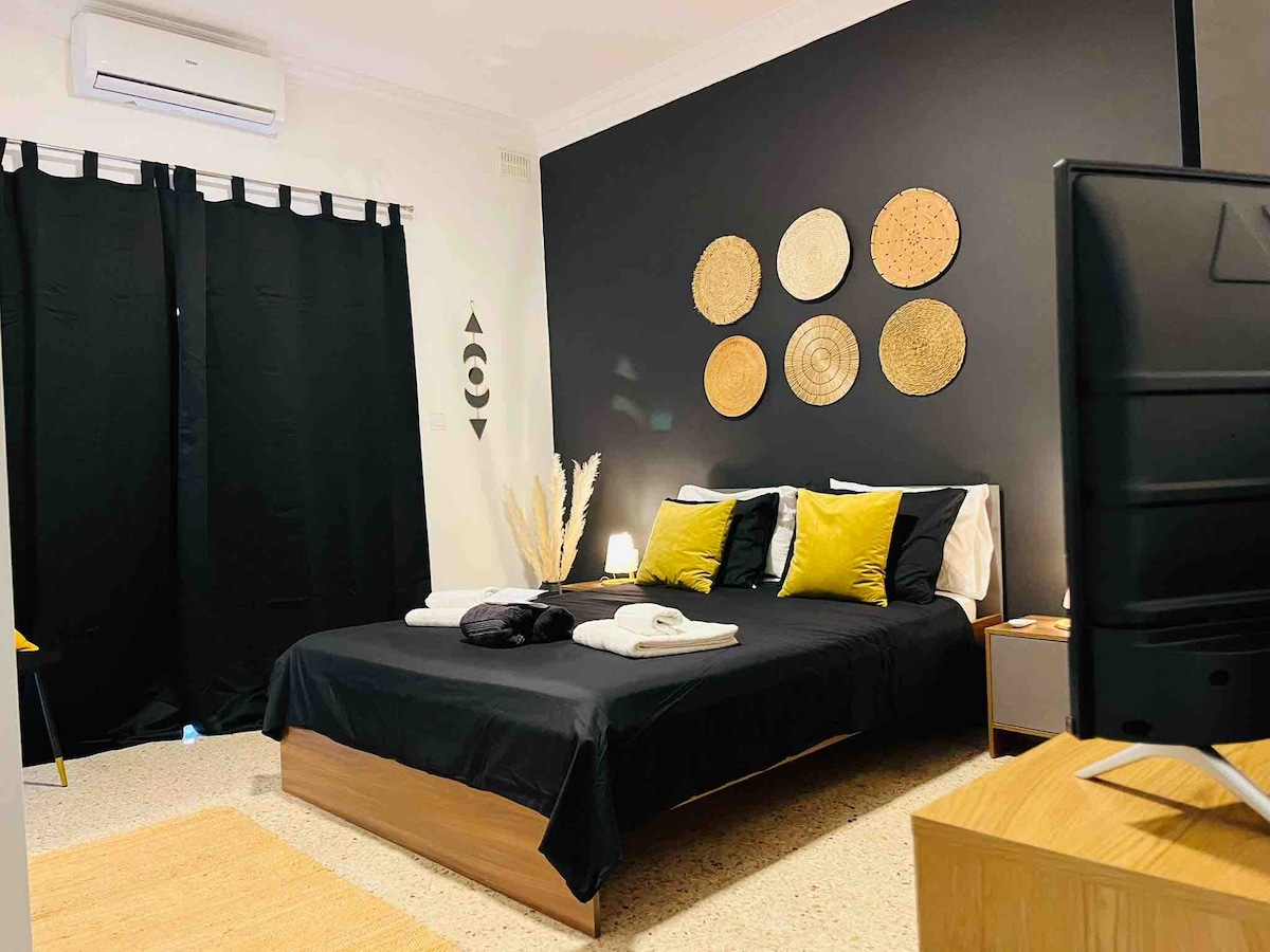A spacious sleeping area features a dark accent wall adorned with decorative plates. The bed, dressed in black linens, is complemented by yellow and black pillows. A bedside lamp and a wooden nightstand add functionality, while black curtains frame a window, enhancing the room's ambiance.
