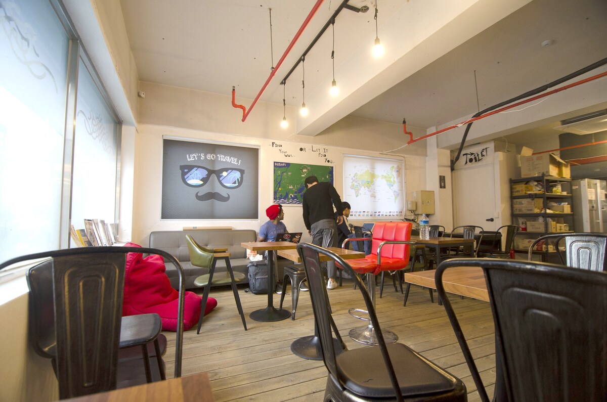 A communal space is presented, featuring a mix of seating options, including chairs and bean bags. Large windows allow natural light to fill the room, which is decorated with travel-themed wall art and a map. A group engages in discussion near a projection screen.