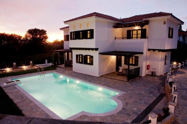 A two-story villa features a spacious outdoor area with a rectangular swimming pool, surrounded by stone flooring. Soft lighting highlights the architecture, while dark green shutters contrast with the white exterior. A serene landscape is visible in the background during sunset.