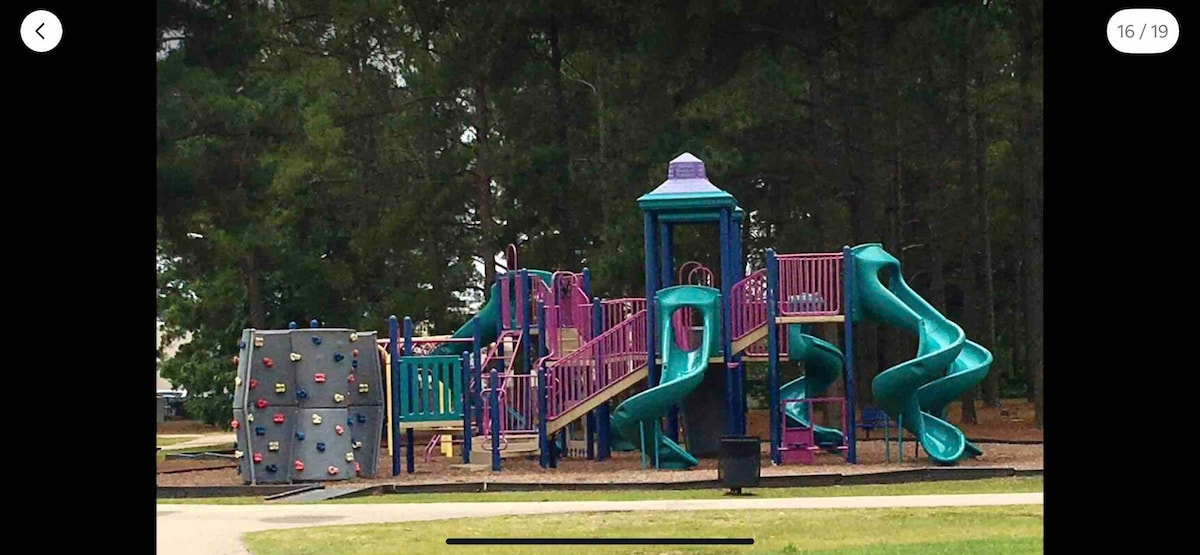 A colorful playground features a climbing wall alongside multiple slides in shades of purple and turquoise. Shaded by trees in the background, the area offers a safe and engaging environment for children to play.