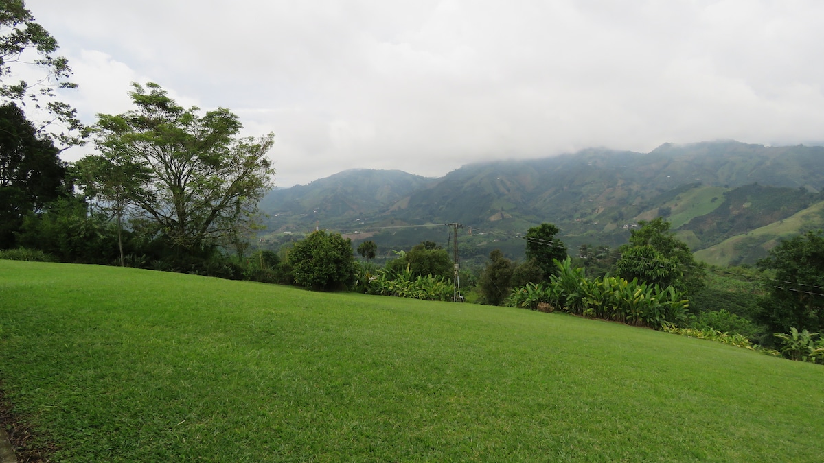 A wide expanse of lush green grass is presented, framed by verdant foliage and trees. The distant mountains are partially obscured by soft clouds, while rolling hills in various shades of green create a serene backdrop, reflecting the natural beauty of the rural landscape.