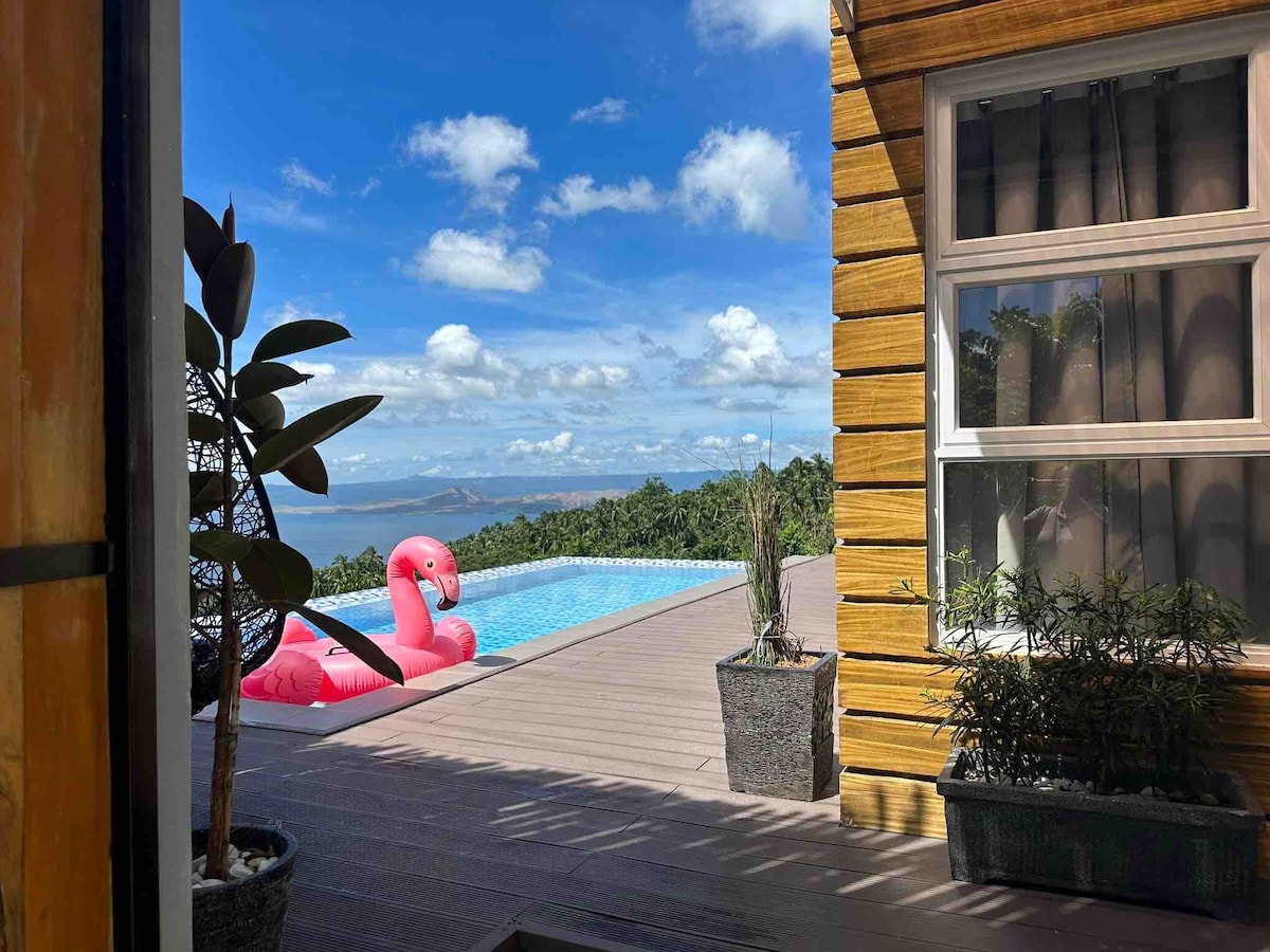 A view from inside the villa reveals an infinity pool surrounded by lush greenery. A pink flamingo float rests in the water, overlooking the scenic landscape of Mt. Maculot and Taal Lake under a clear blue sky.