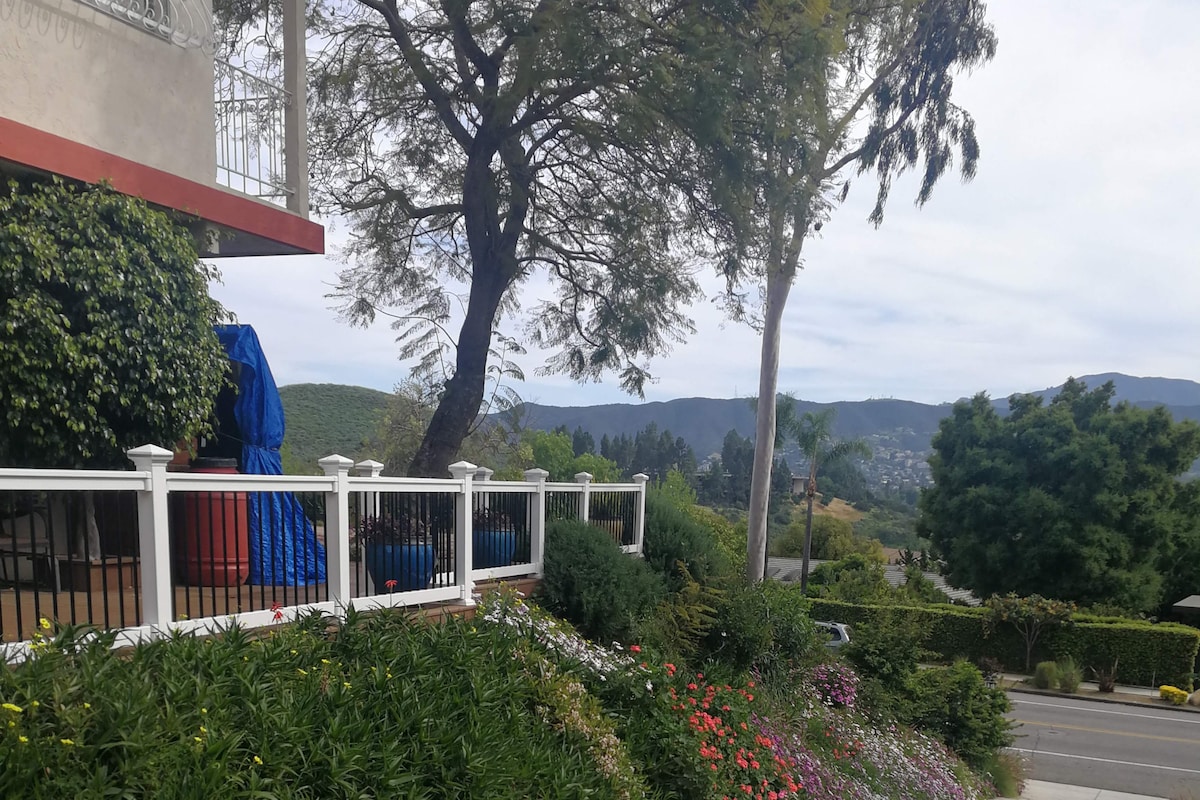 A landscaped outdoor area features a white picket fence with various shrubs and flowers in bloom. A tree stands nearby, with a scenic view of the mountains and hills in the background under a lightly clouded sky.