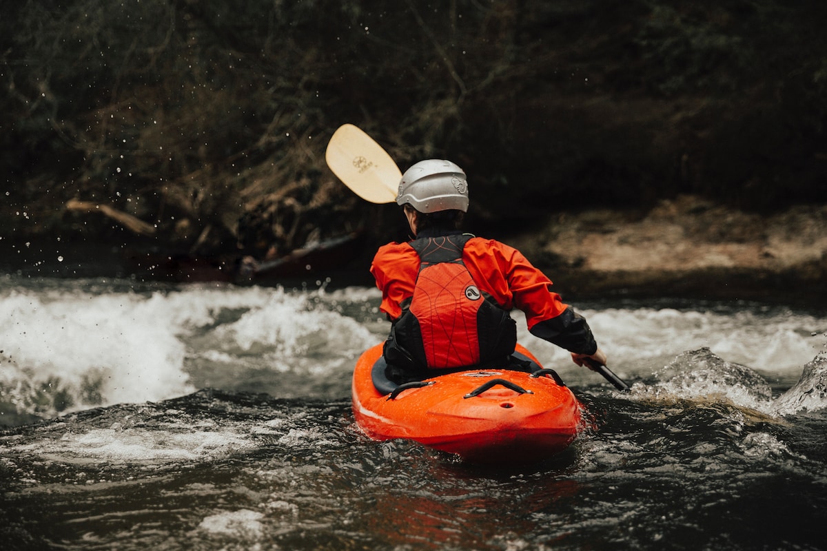 A person wearing a red kayak suit and helmet is paddling an orange kayak through swirling waters. Surrounding trees create a natural backdrop. The kayak's bow is directed toward the rushing water, indicating movement and engagement with the river's current.