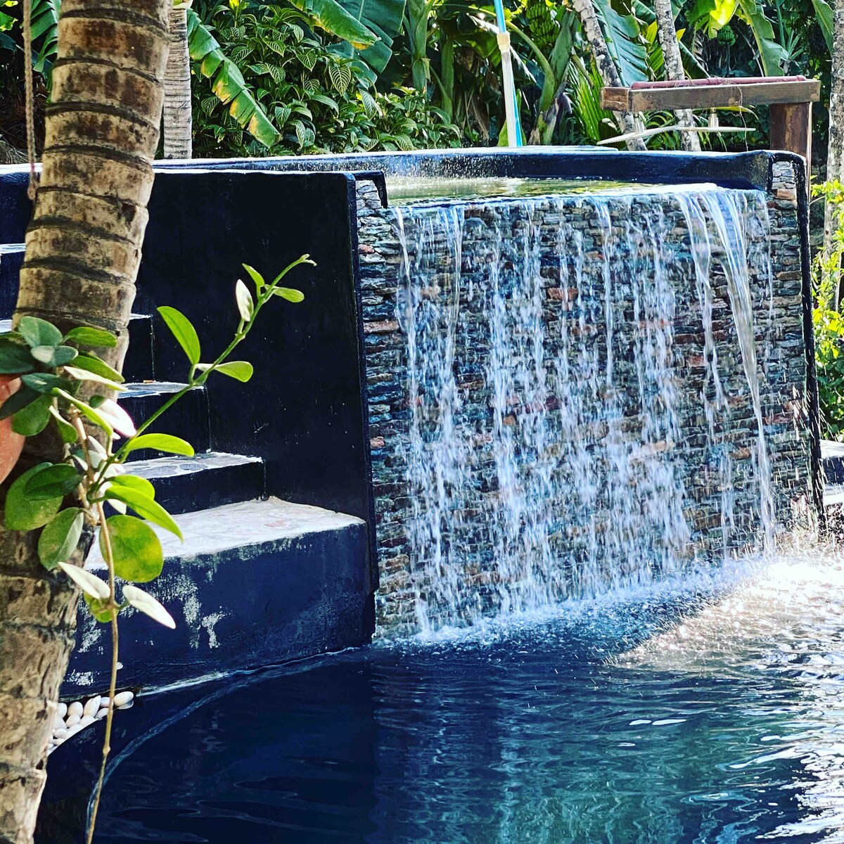 A serene waterfall cascades over natural stone, creating a soothing ambiance. The pool, surrounded by lush tropical foliage, invites relaxation. Steps lead gracefully from the water's edge, enhancing accessibility to this tranquil oasis.