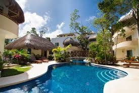 A serene outdoor pool area is surrounded by lush greenery and tropical plants. The pool features clear blue water, with a gently curving shape. Thatched-roof structures and nearby lounging areas provide shaded spots for relaxation in this tranquil environment.