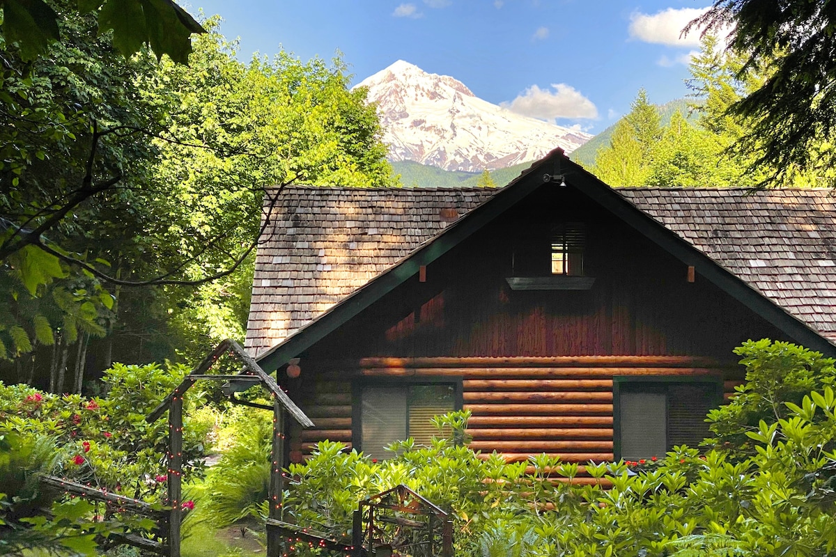 A rustic log cabin is nestled among lush greenery, with vibrant foliage framing the entrance. Mt. Hood rises majestically in the background, providing a stunning contrast to the wood exterior and the blue sky above.