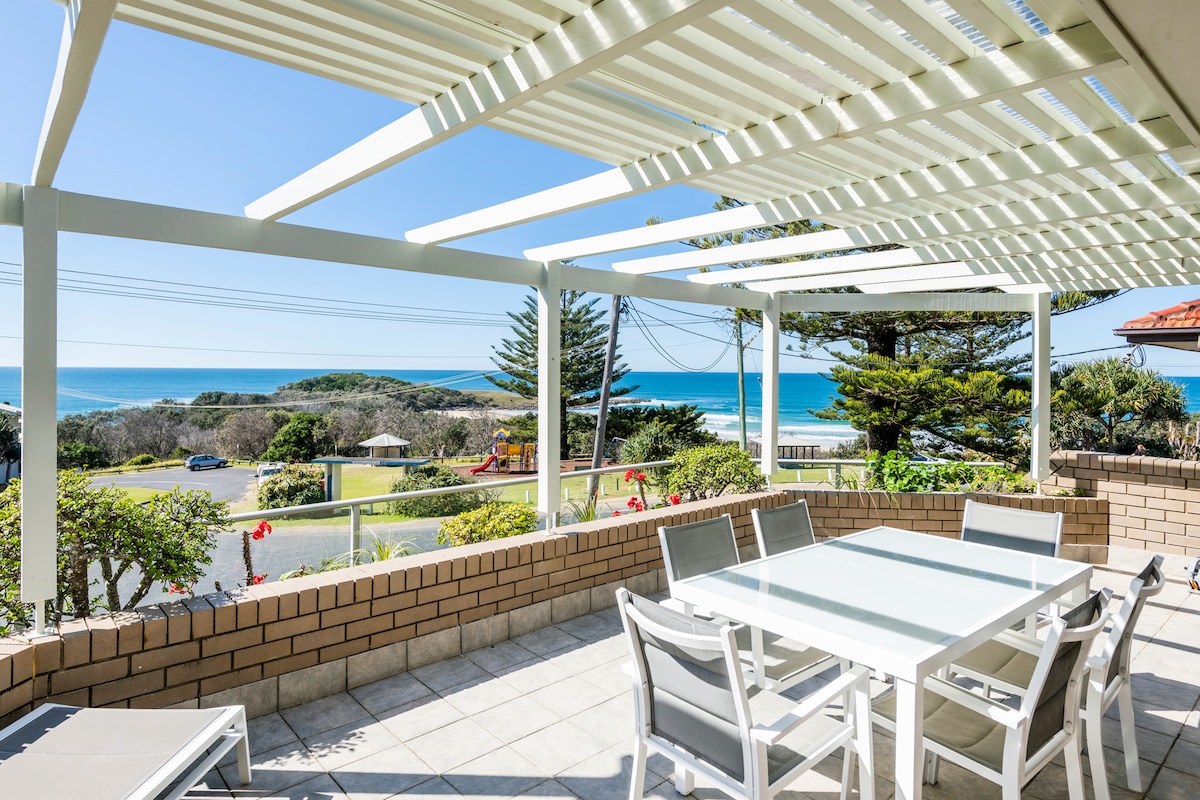 An outdoor deck area is featured, furnished with a dining table and several chairs. A pergola overhead provides shade. The expansive view overlooks the Pacific Ocean and greenery, with the shoreline visible in the distance under clear blue skies.