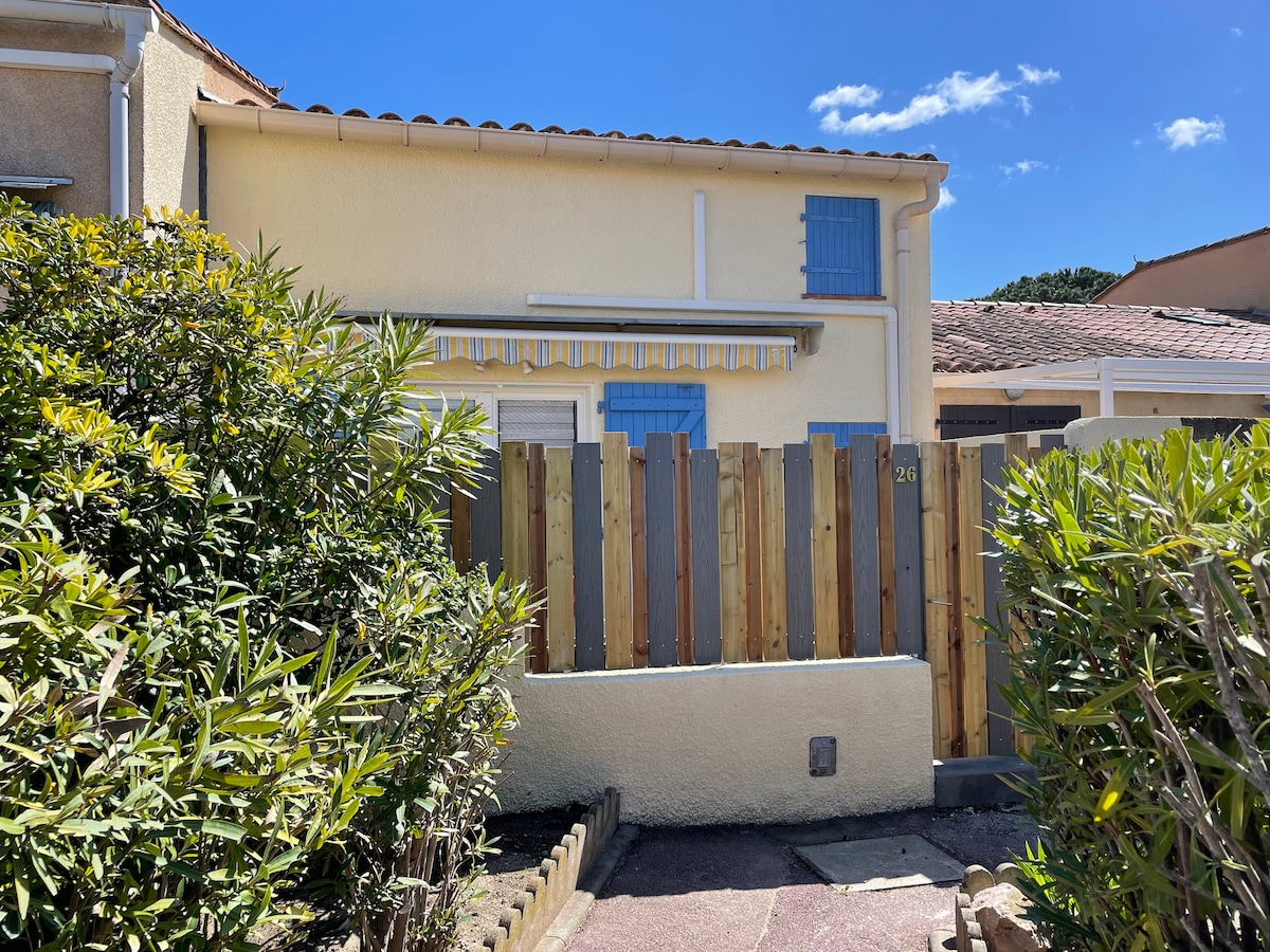 A charming exterior of the maisonette is featured, showcasing light-colored walls framed by blue shutters. A wooden fence provides privacy, while vibrant greenery surrounds the entrance, creating a welcoming environment. The clear blue sky above enhances the inviting atmosphere.