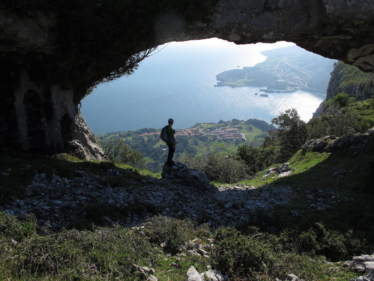 A person stands at the edge of a rocky opening, overlooking a scenic coastal view. The ocean shimmers in the sunlight and small villages are visible down below, framed by lush greenery and rugged cliffs.