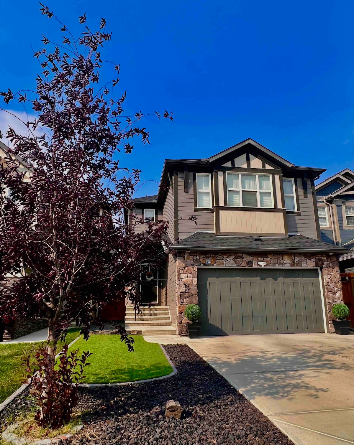 A two-story house is presented in the image, featuring a combination of stone and siding on the exterior. A well-maintained flowerbed with dark mulch surrounds the base. A contemporary garage door is visible, with stairs leading to the entrance. A small tree with purple foliage stands in the front yard.