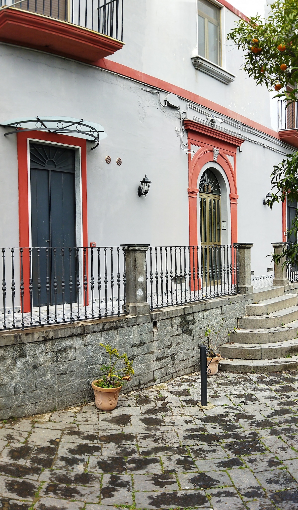 The entrance of a historic villa is framed by a red-trimmed archway and a decorative metal lantern. Stone steps lead up to the door, flanked by a wrought iron railing. Potted plants add a touch of greenery along the pathway.