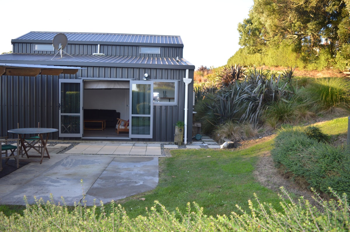 The exterior shows a modern studio with a metal façade, featuring a spacious patio area. A set of glass doors opens to the living space. The surrounding landscape includes well-maintained grass and native plants, contributing to a serene environment.