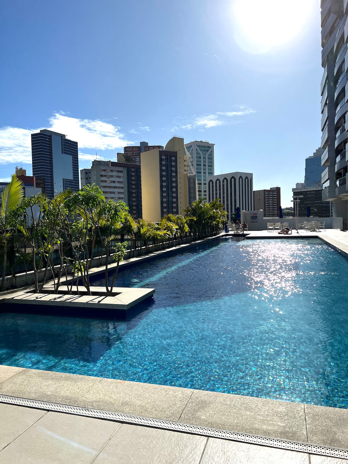 A large outdoor pool reflects the clear sky, surrounded by palm trees and modern buildings in the background. The pool area features a spacious deck for relaxation, enhancing the overall atmosphere of comfort and leisure.