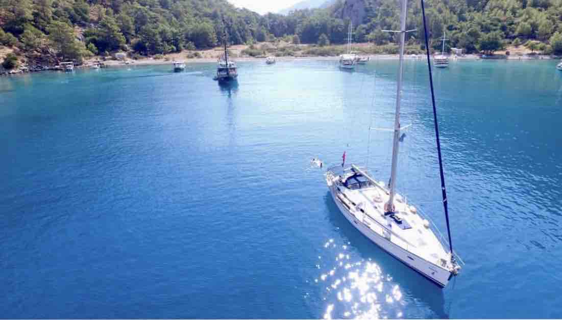 A sailboat is anchored in calm, turquoise waters surrounded by lush greenery and distant hills. The clear, serene water reflects sunlight, creating shimmering patterns. Additional boats can be seen in the background, adding to the tranquil maritime atmosphere.