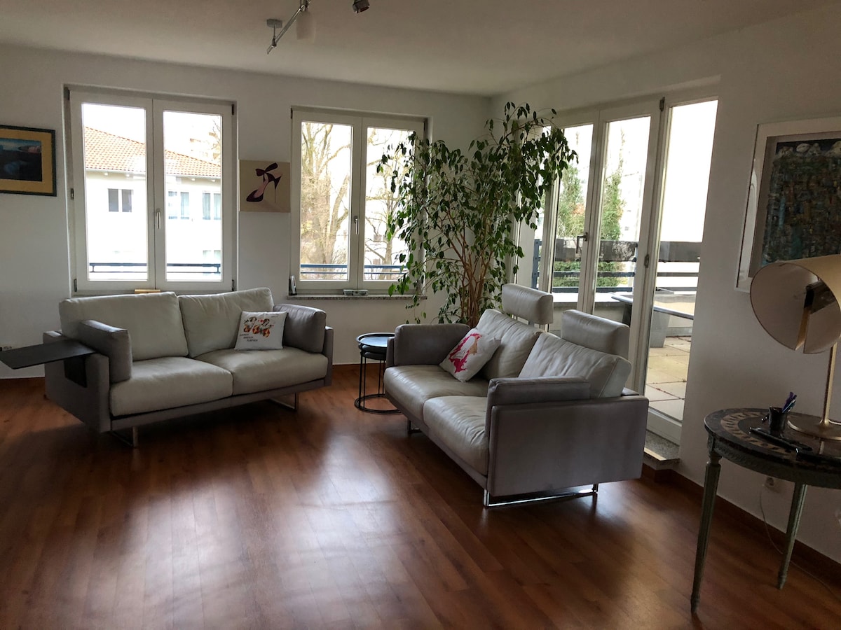 A spacious living area is presented with large windows allowing natural light to fill the room. Two modern sofas in light grey are positioned for comfort, accompanied by a small round table. A decorative plant adds a touch of greenery, enhancing the inviting ambiance.
