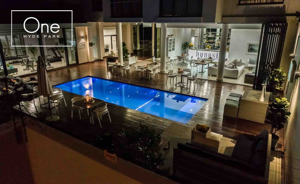 An outdoor pool area is highlighted by a shimmering blue swimming pool surrounded by lounging furniture. Ambient lighting illuminates the space, enhancing a serene environment. Nearby, dining tables and chairs are arranged, creating an inviting area for relaxation and socializing.