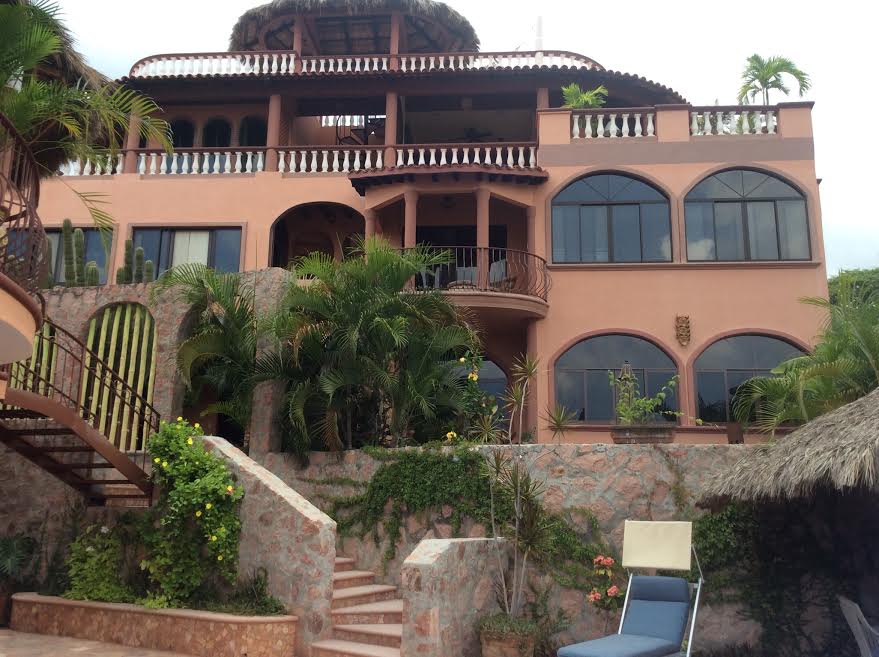The exterior of the villa displays a multi-tiered design with a thatched roof. Large arched windows reflect the surrounding greenery. A stone pathway leads down to the pool area, while tropical plants frame the entrance, enhancing the natural beauty of the property.