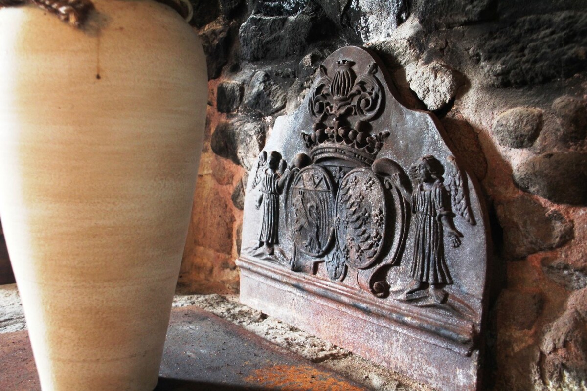 An ornate stone fireplace is featured with intricate carvings portraying figures and crests. A simple ceramic vase is placed nearby, adding a touch of rustic charm to the setting. The stone wall reflects a traditional and historic design.