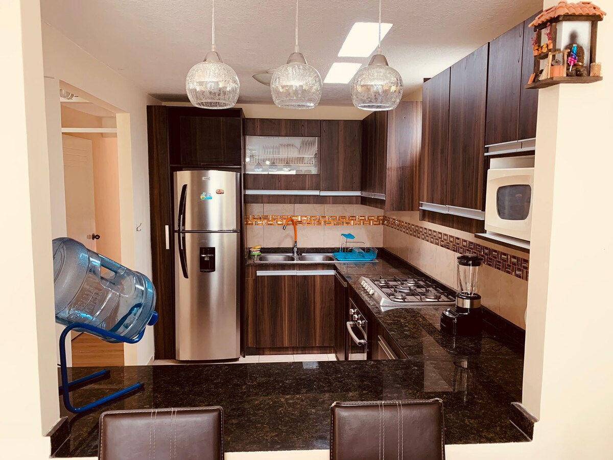 A modern kitchen features dark wooden cabinetry, stainless steel appliances, and a black granite countertop. Hanging pendant lights provide illumination above the kitchen island. A water dispenser is present, contributing to convenience within the space.