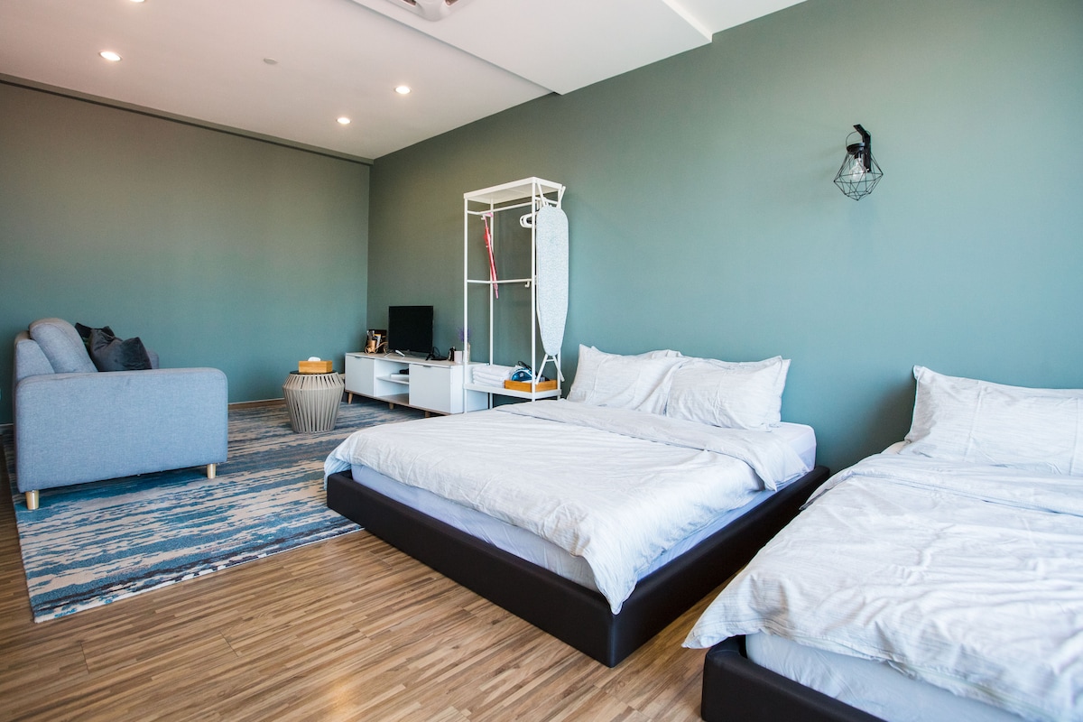 A spacious room features two queen beds covered with white linens, complemented by a soft blue area rug. A three-seater couch and a TV stand are positioned against the green wall, providing a relaxed atmosphere. Natural light is available through ceiling lights.