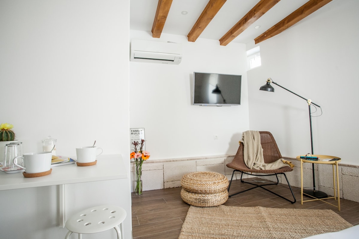 The room features a light color scheme with wooden beams across the ceiling. A comfortable chair is positioned beside a small table, which holds cups and a teapot. A wall-mounted TV is visible, and a cozy throw drapes over the chair.