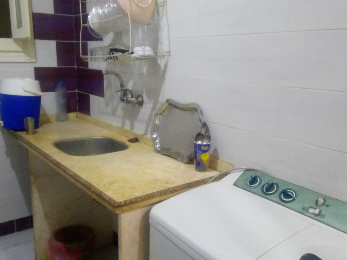 A functional kitchen area is captured, featuring a small sink, countertop, and a washing machine. Clean dishes are neatly arranged on a wall-mounted rack. A tray and a cup are also visible on the counter, with a blue container located nearby.