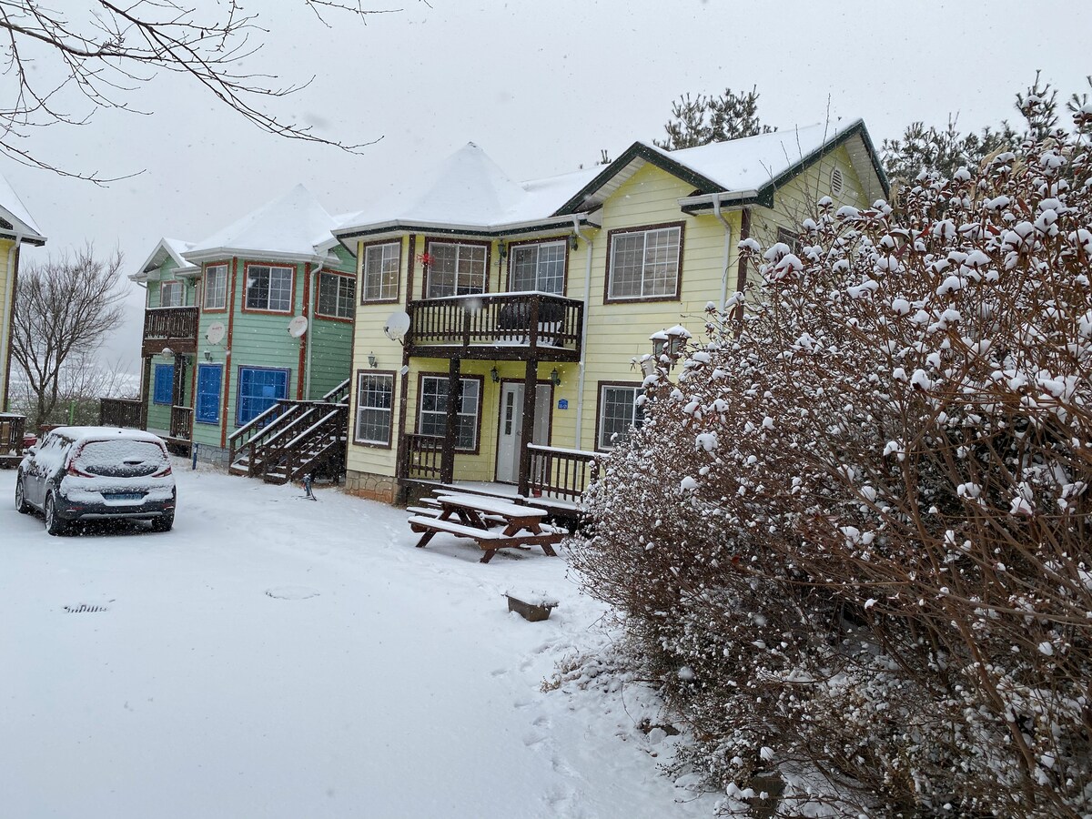 A charming multi-colored building stands in a snowy landscape, showcasing a mix of green and yellow facades with multiple balconies. A picnic table is set outside, along with a parked car. Snow blankets the ground, enhancing the serene atmosphere of the surroundings.