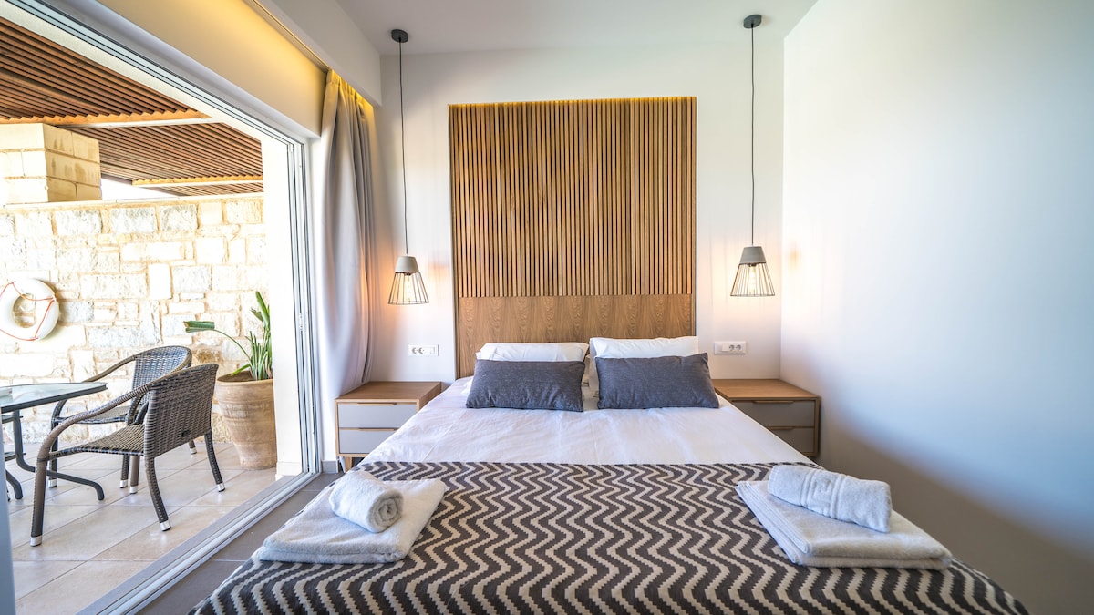 A spacious bedroom features a wooden accent wall behind a large bed, complemented by two bedside lamps. Light grey linens are layered with a patterned blanket. A terrace is visible through the sliding glass door, revealing outdoor seating and greenery.