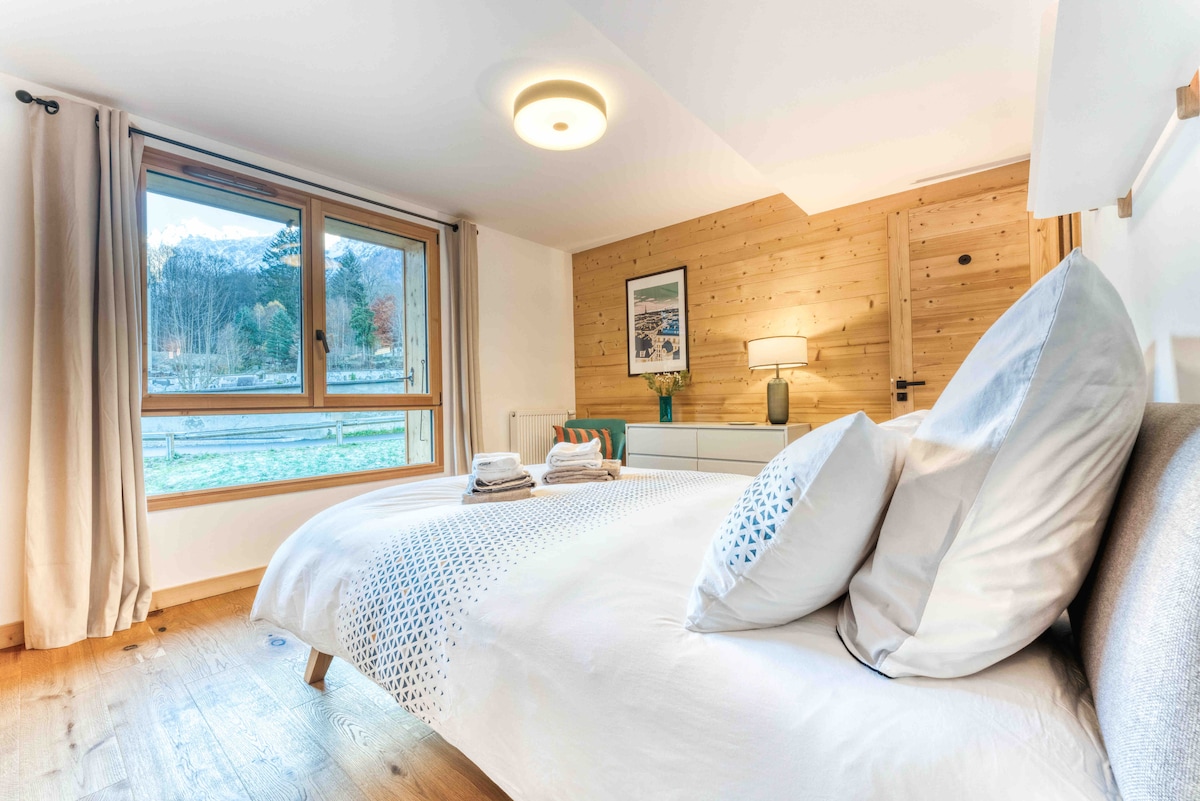 A serene bedroom features a spacious bed dressed with soft linens and plump pillows. Natural light floods the room through a large window, offering views of the surrounding landscape. A light wood accent wall and tasteful decor elements create a calming atmosphere.