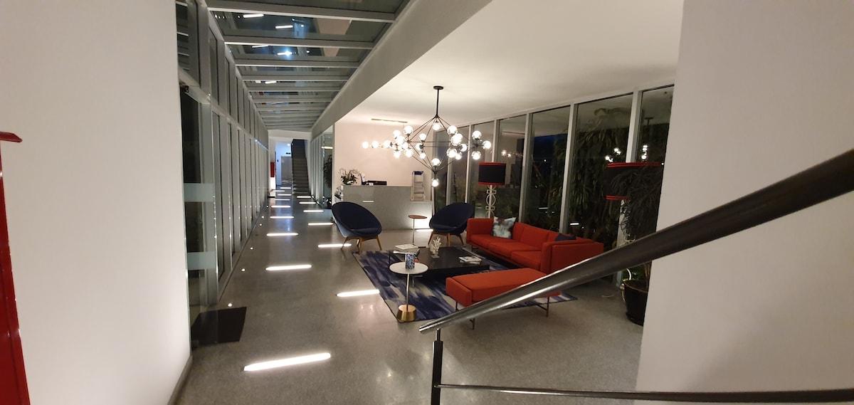 A modern lobby area is presented, featuring a combination of sleek furnishings and large windows. A striking lighting fixture hangs above a seating arrangement, which includes blue chairs and an orange sofa. The space is illuminated by soft overhead lights along a polished floor.