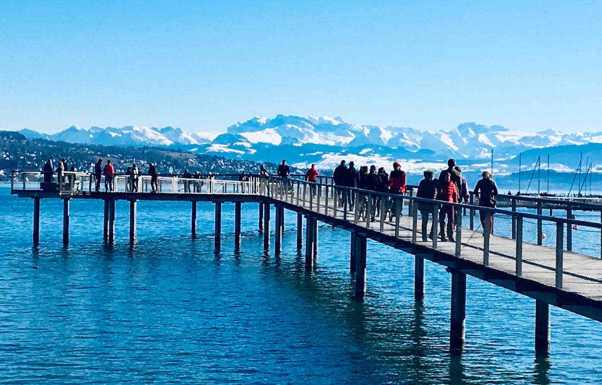 A wooden pier extends into clear blue water, with people walking and enjoying the view. Snow-capped mountains create a stunning backdrop against a bright sky. The pier features a sturdy design with railings, allowing for safe access to the waterfront.