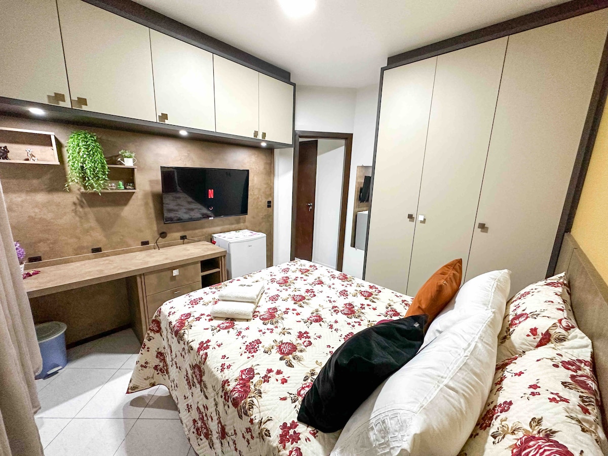 A comfortably arranged room features a spacious bed adorned with floral bedding and several pillows. A wall-mounted Smart TV is visible above a simple desk, accompanied by a minibar. Storage cabinets line the upper wall, and a touch of greenery is seen on a shelf.