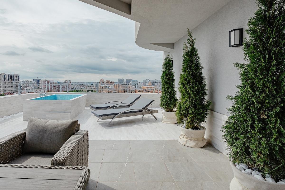 A spacious terrace offers a view of the city skyline and distant hills. Two stylish lounge chairs are positioned beside a mini-pool, while potted trees frame the area. The surface is tiled with light-colored stone, enhancing the modern aesthetic.