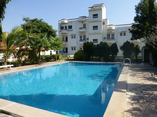 A large swimming pool surrounded by lush greenery is showcased. The clear blue water reflects the sunlight, inviting guests to relax. A white building with multiple windows stands in the background, providing a serene setting for leisure and recreation.