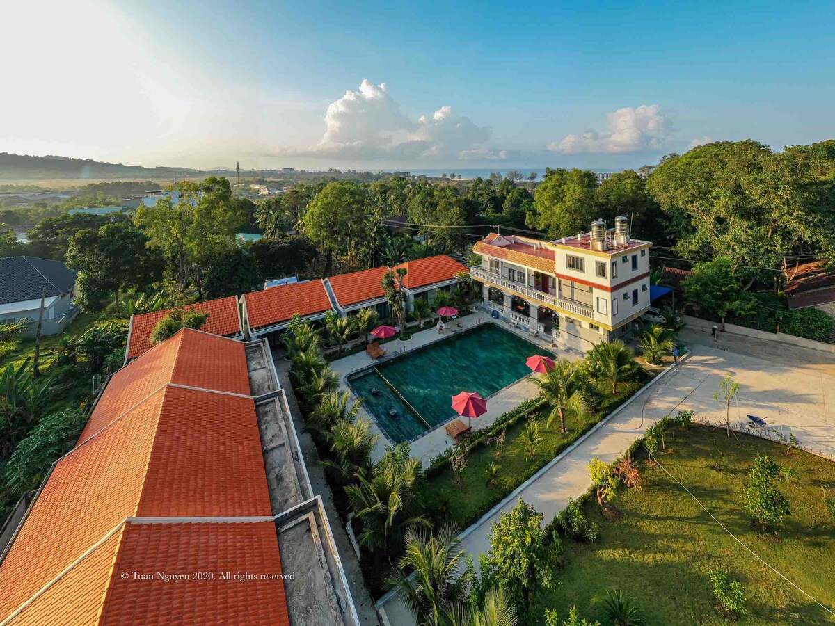 Aerial view of the bungalow complex highlights a central swimming pool surrounded by lush greenery. Red umbrellas provide shade for outdoor seating areas. The terracotta roofs of the buildings contrast with the verdant landscape, while the horizon reveals the distant sea.