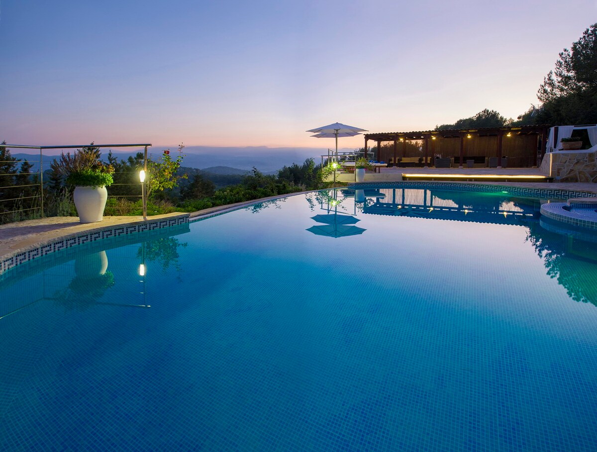 The serene pool reflects the evening sky, surrounded by lush greenery and softly illuminated landscaping. A shaded lounge area is visible in the background, complemented by an umbrella. The expansive view of the horizon adds to the tranquil setting.