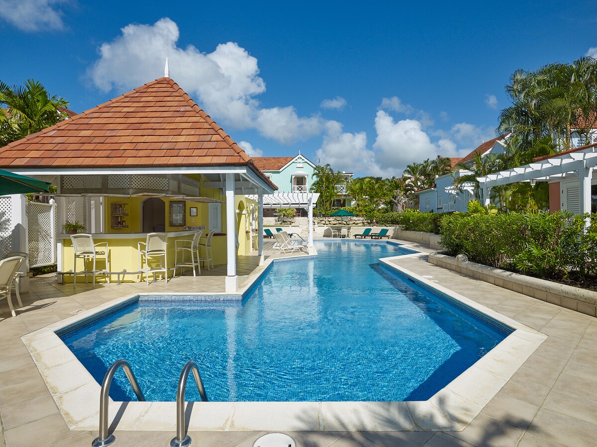 A clear blue swimming pool is framed by a sunlit patio, featuring a small attached gazebo with a bar area. Lush greenery and vibrant tropical plants border the pool, contributing to a serene environment. Sun loungers are arranged around the pool, inviting relaxation.