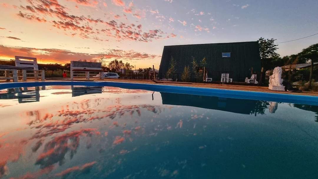 The image captures a tranquil swimming pool surrounded by a peaceful rural landscape. The clear water reflects vibrant colors from the sunset sky, while a simple cabin structure stands in the background, accompanied by minimal outdoor seating and greenery.