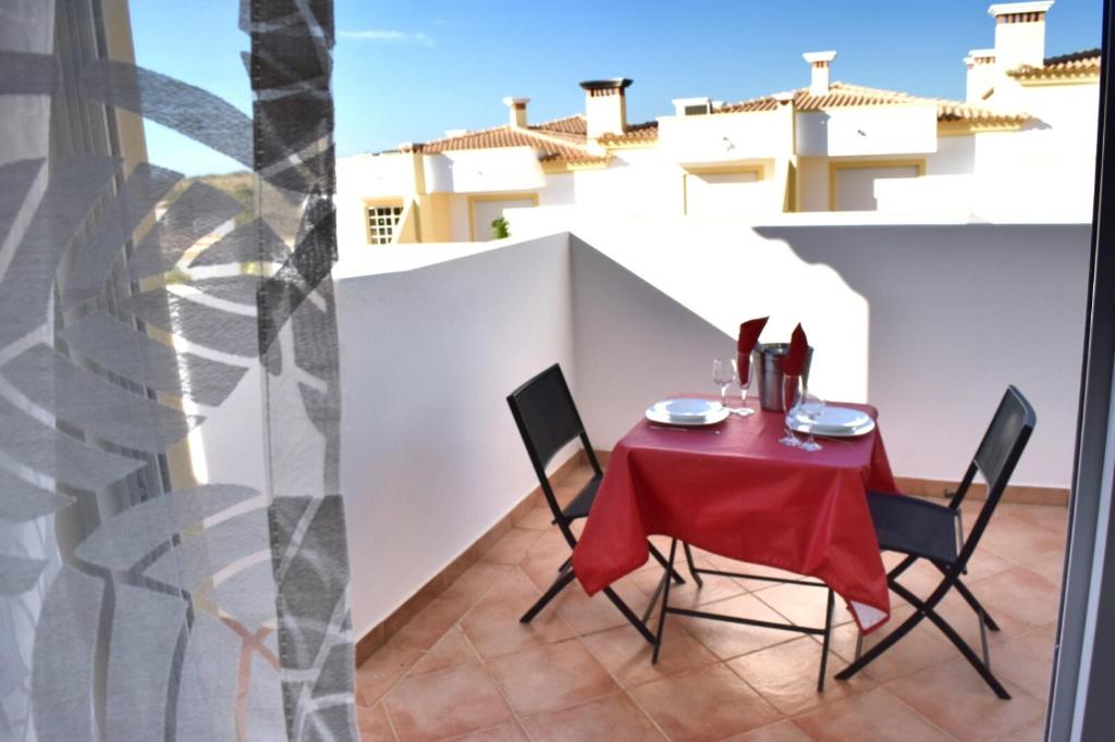 An outdoor dining area features a round table set for two, draped in a red tablecloth. Two black chairs are positioned around the table, with a scenic view of the neighborhood rooftops visible in the background. Soft natural light enhances the inviting space.