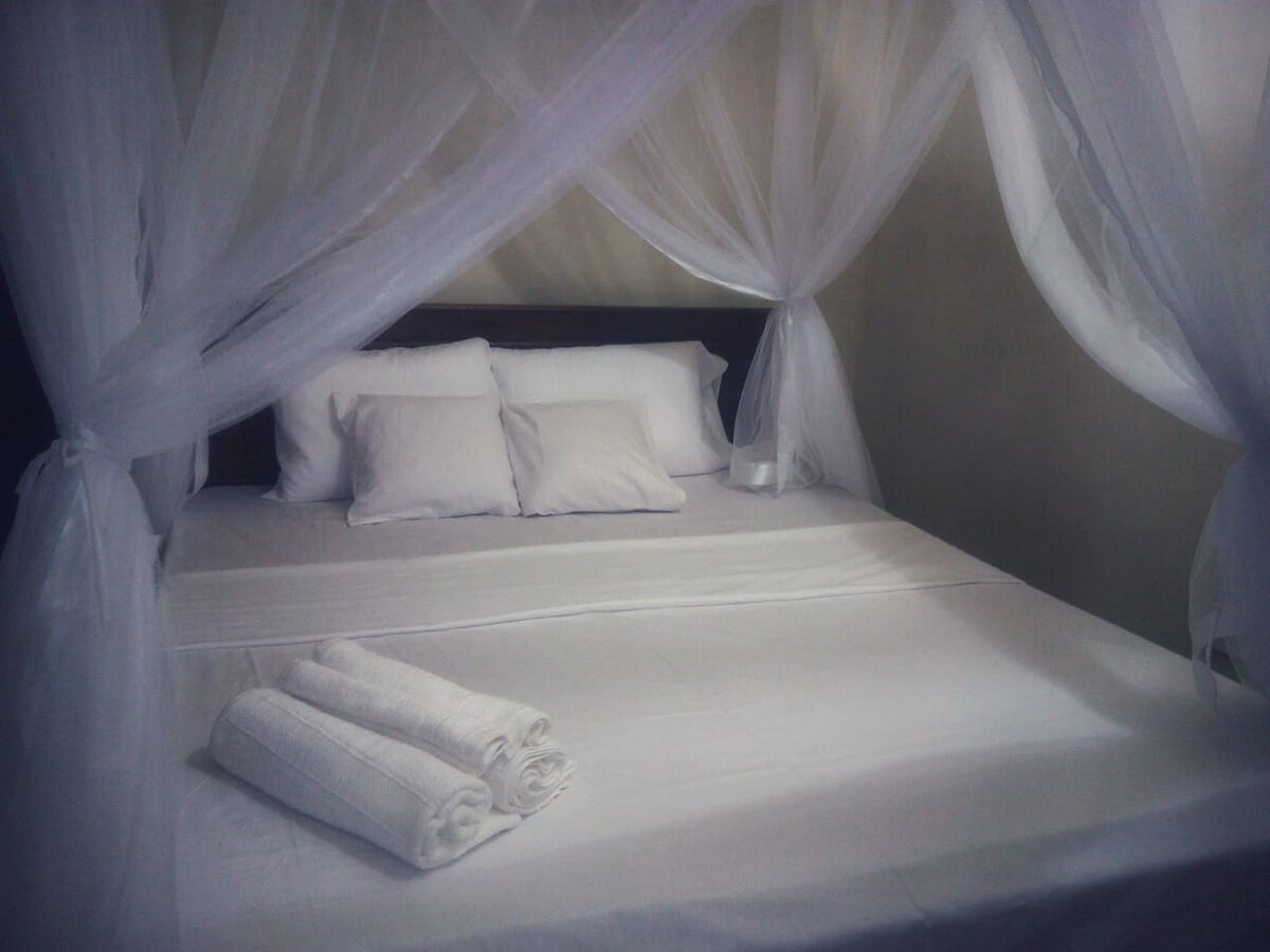 A spacious bed is adorned with a white canopy, creating a gentle, airy feel. Fresh towels are neatly arranged at the foot of the bed, while soft, neutral-toned linens provide a calming backdrop.