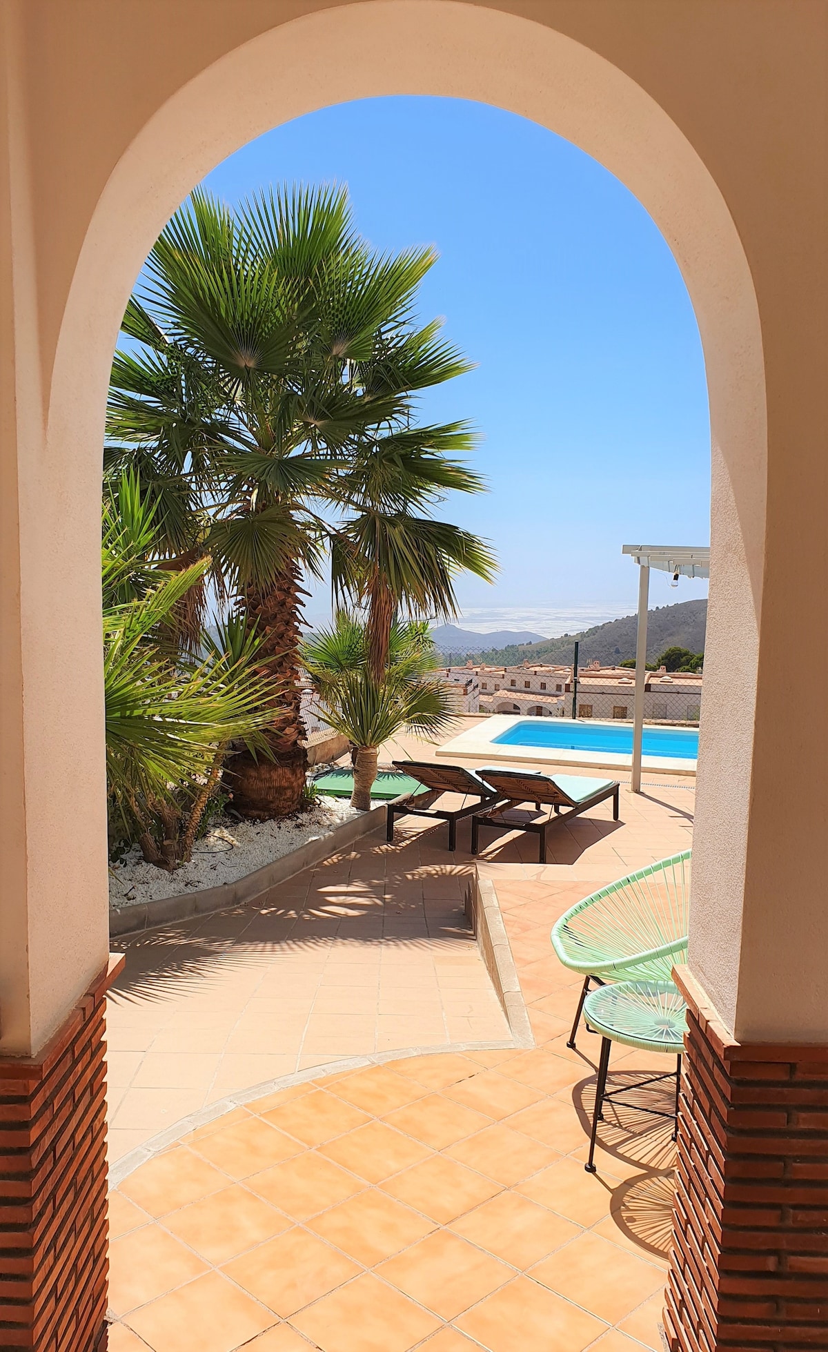 An archway frames a view of the outdoor area, featuring palm trees and a private pool. Sun loungers and a table are positioned on a tiled patio, with hills visible in the distance under a clear blue sky.