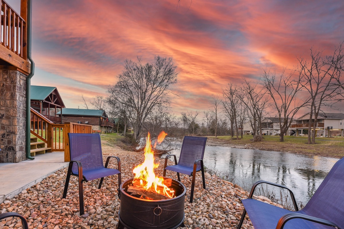 A fire pit is surrounded by four blue chairs, set on pebbles beside a calm river. The sky is painted in vivid sunset hues of orange and purple, while trees line the riverbank, creating a peaceful outdoor setting.