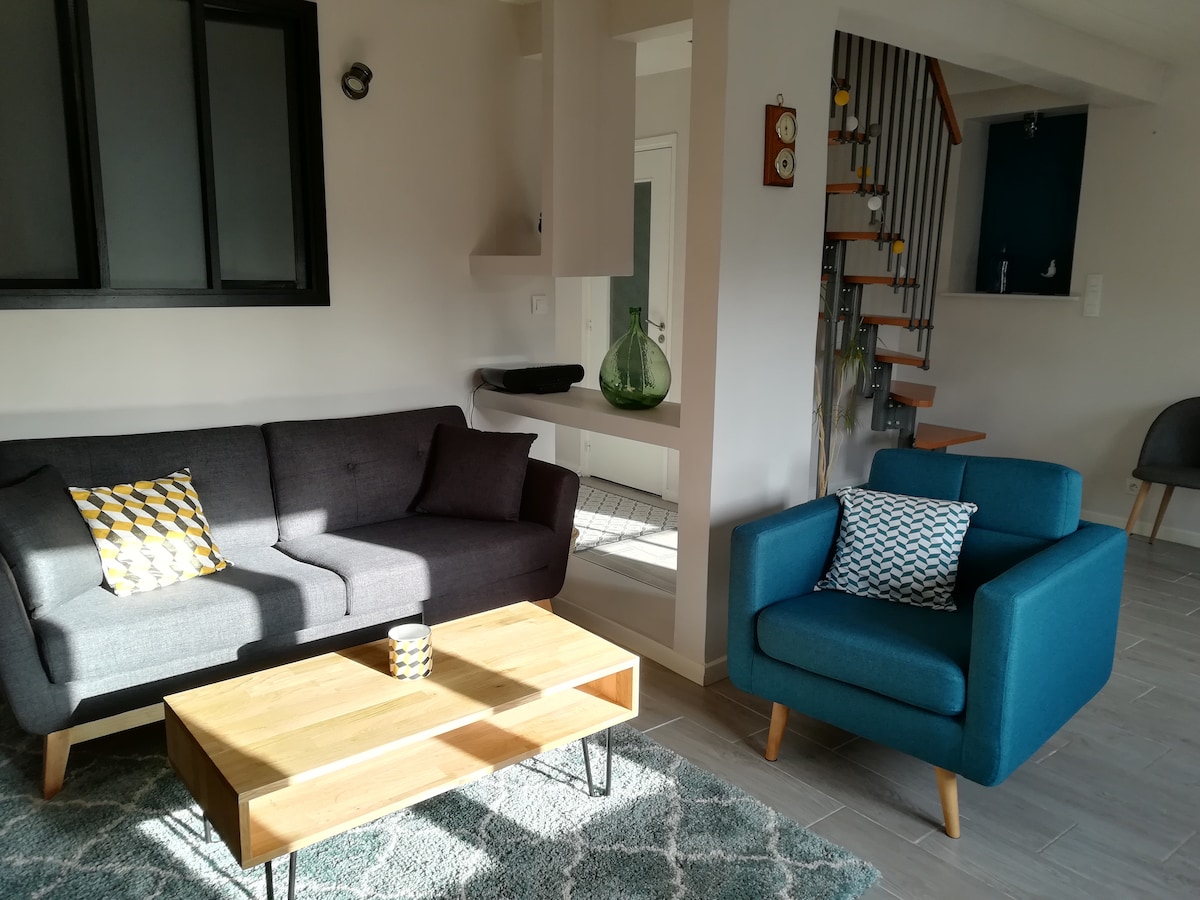 A living room area features a grey sofa with a patterned cushion and a blue armchair with a decorative pillow. A wooden coffee table sits in front, and light filters in through large windows, enhancing the airy atmosphere. A staircase is visible in the background.
