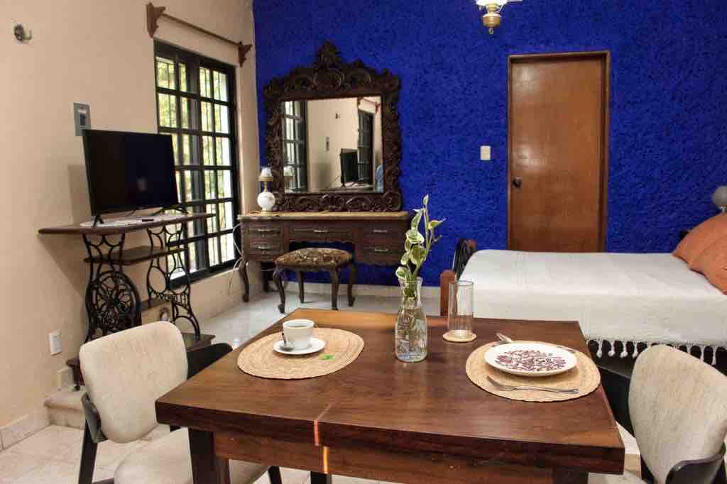 A cozy living area features a wooden dining table set with decorative placemats, a vase holding a single flower, and a cup. A carved wooden mirror complements the vibrant blue wall. A television and a spacious bed are visible, enhancing the room's vintage charm.