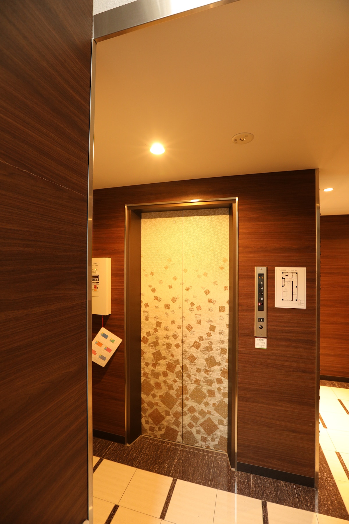 An elevator entrance is framed by warm wooden panels. The elevator doors are adorned with a subtle pattern, and a control panel is mounted beside the entrance. Soft lighting illuminates the space, enhancing the clean and modern aesthetic of the building's interior.