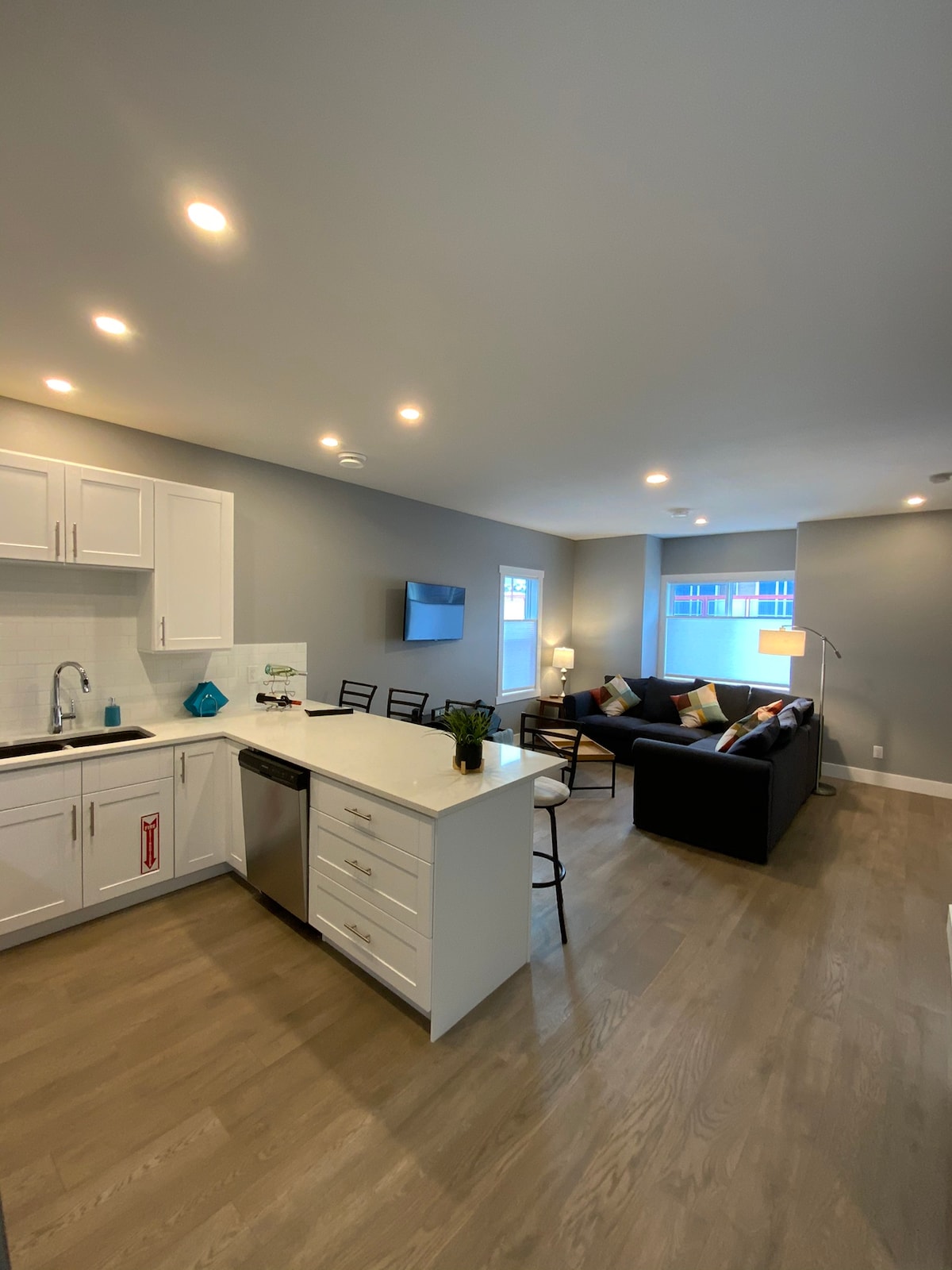 An open-plan living area features modern white cabinetry and a spacious kitchen island with seating for two. Natural light enters through the window, illuminating a comfortable seating area with a black couch, decorative pillows, and a wall-mounted TV. Clean lines and neutral colors define the space.
