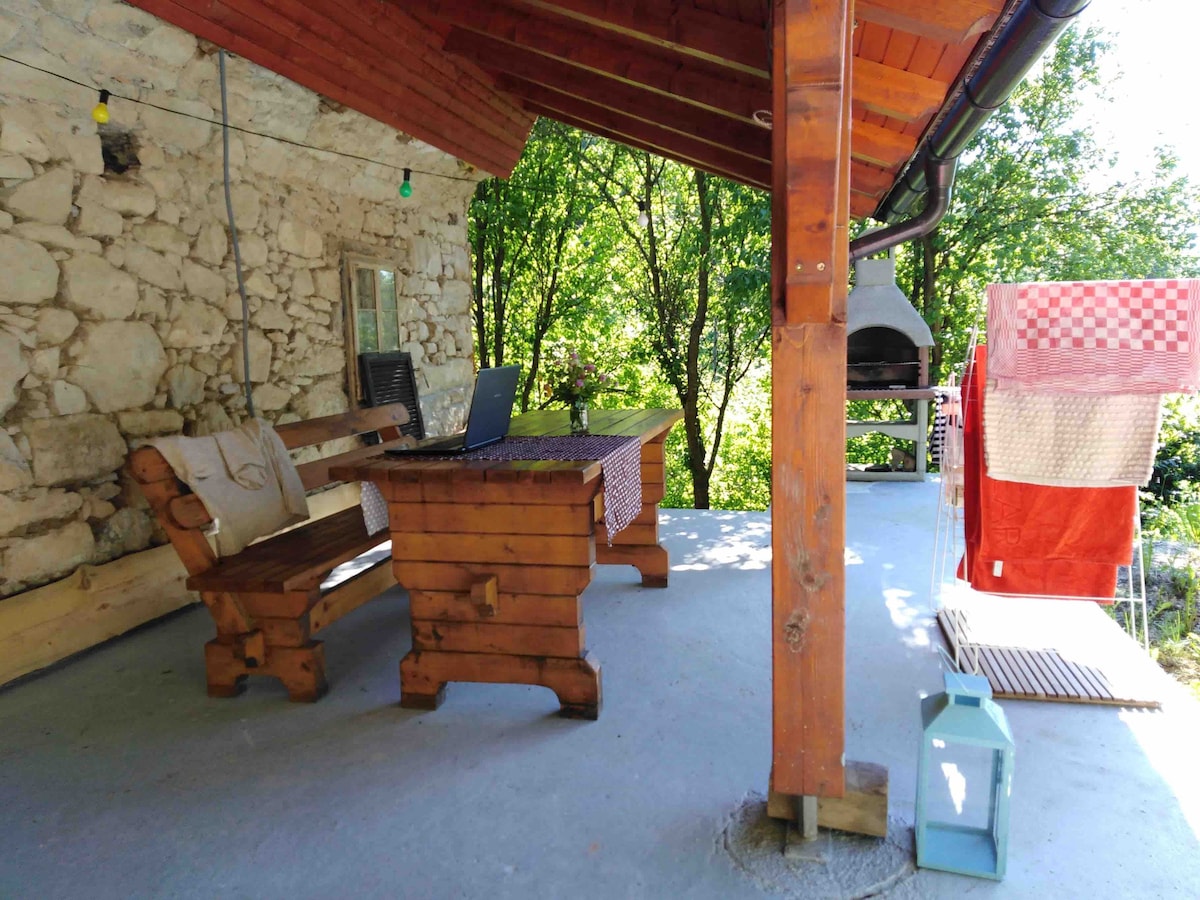 An outdoor terrace features a wooden table and bench set, surrounded by trees. A laptop is placed on the table, with a barbecue grill seen in the background. Wash lines with colorful items are positioned to the side, adding a casual touch to the space.